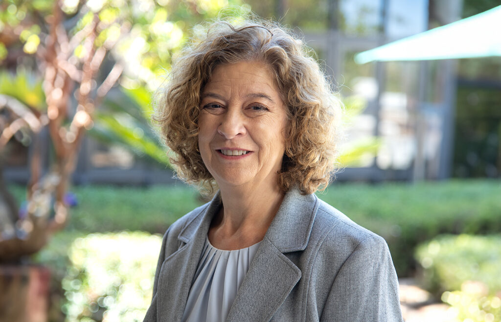 Allison Baird-James smiling in a gray blazer, outdoors with greenery in the background