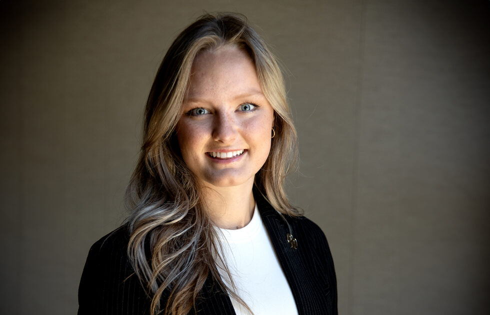 Ashleigh Spragins smiling in a dark jacket with long blonde hair, standing in soft lighting