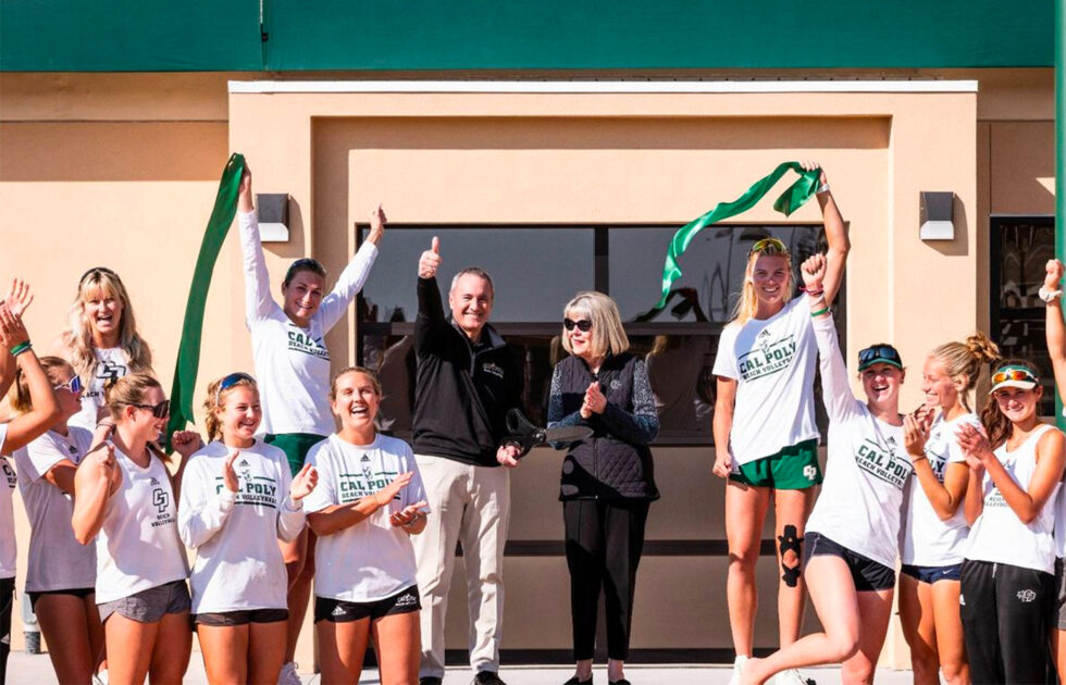 Group of Cal Poly beach volleyball players and staff celebrating with a ribbon cutting in front of the new clubhouse