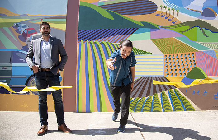 Two people at a ribbon-cutting ceremony in front of a vibrant mural depicting agricultural and coastal landscapes