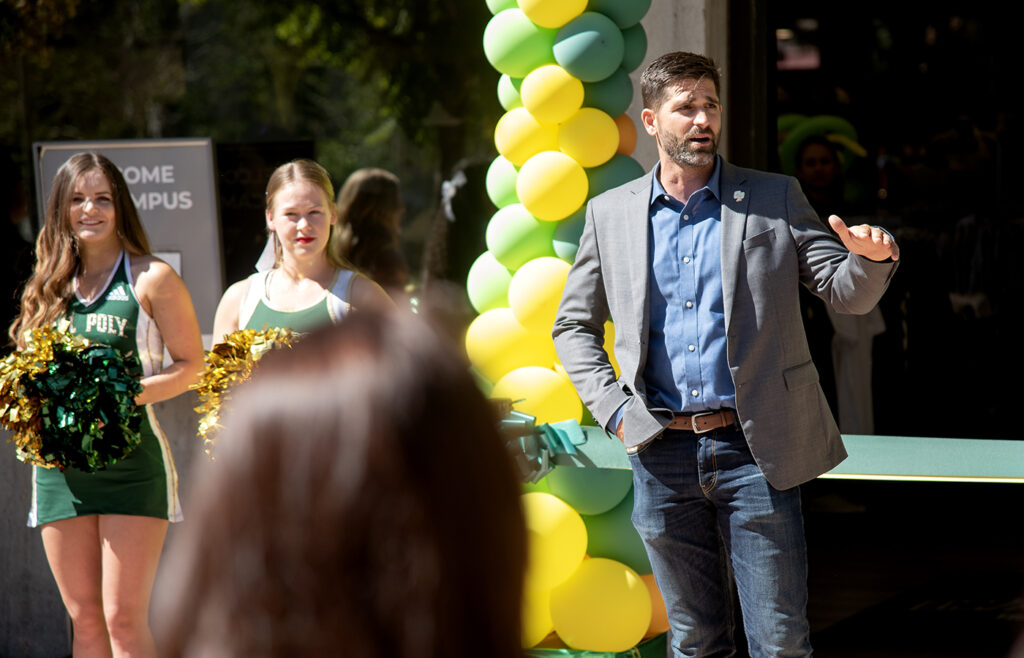 Cal Poly staff member speaking at ribbon-cutting ceremony with cheerleaders and green-yellow balloons