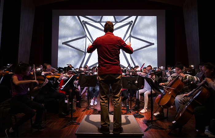 Conductor leading a Cal Poly symphony performance with a geometric visual projected behind the orchestra