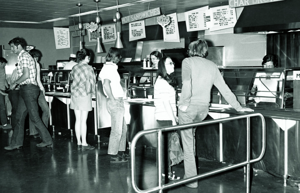Students standing in line at a 1960s-era university dining hall with stainless steel food stations and menu boards overhead.