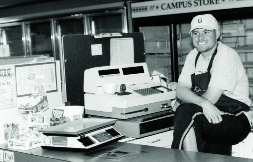 Campus store employee in the 1970s sitting by a register with snacks and a scale on the counter.