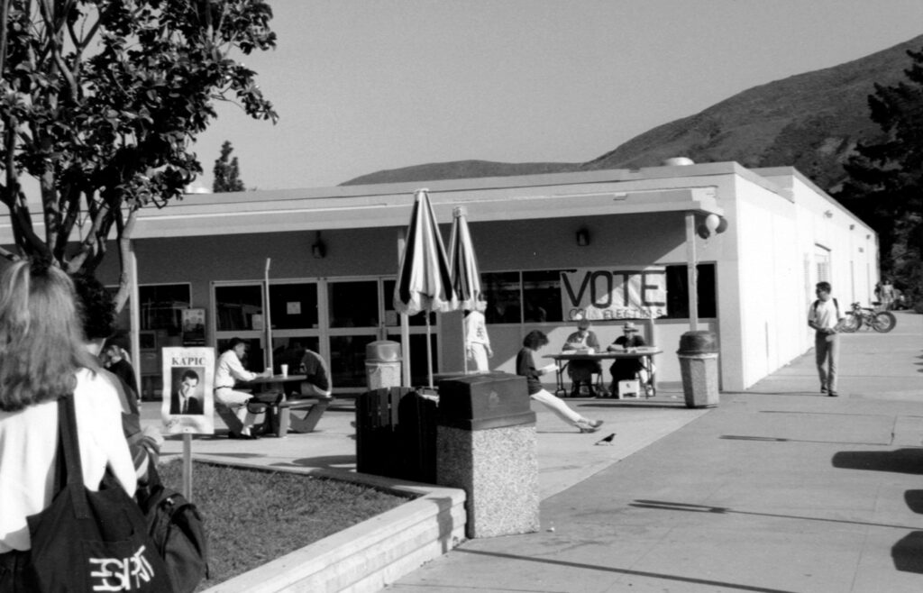 Black and white photo of students outside a campus building during ASI elections in the 1980s, with a 'VOTE' sign visible.