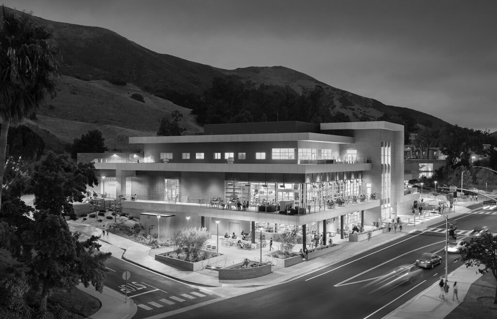 Evening view of the modern Cal Poly dining complex with illuminated windows, hillside backdrop, and students walking outside.
