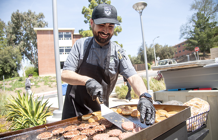 Campus Dining chef smiling while grilling burgers and buns outdoors on a sunny day