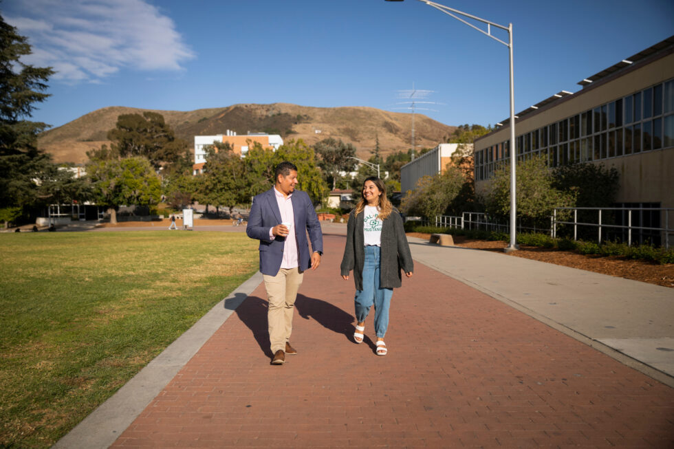Two people walking and talking on a university campus pathway with hills in the background
