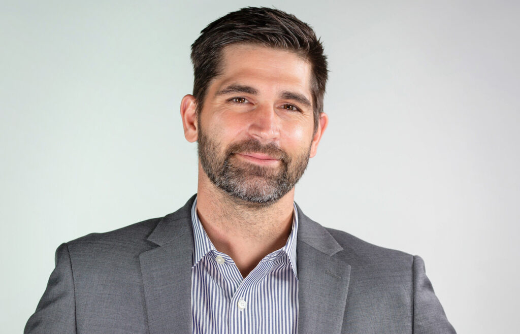 Portrait of Cody in a gray suit jacket and striped shirt, smiling against a plain light background