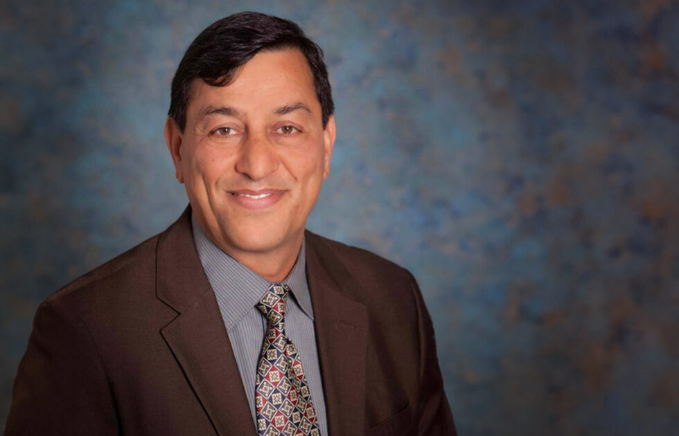Cyrus Ramezani in a brown suit and patterned tie smiling in front of a mottled blue background