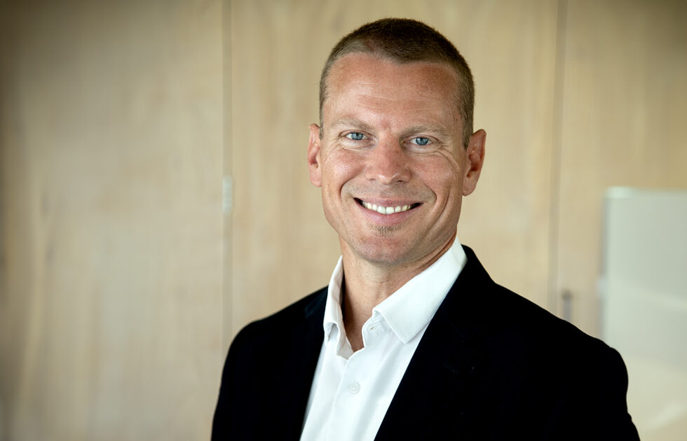 Damon Fleming smiling in a black jacket and white shirt, in a light wooden office setting