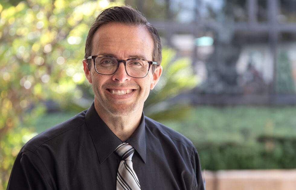 Professional headshot of Dan Banfield in a black shirt and striped tie with green plants in the background
