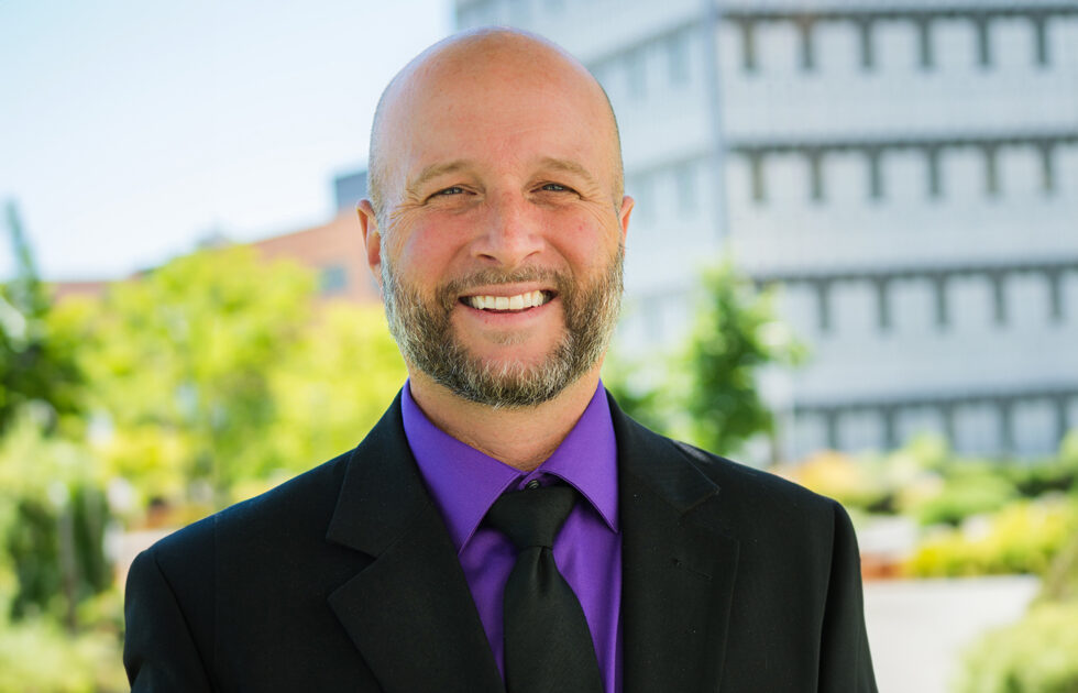 Dean Wendt smiling in a black suit and purple shirt with a black tie, outdoors with campus buildings in background