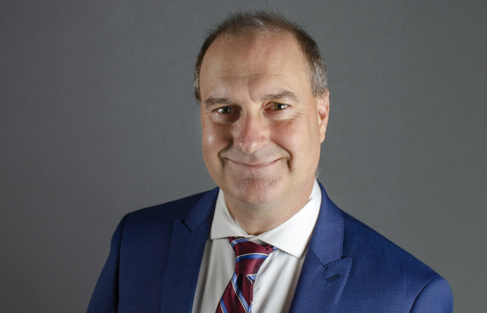 Sean Hurley wearing a blue suit and red striped tie smiling in front of a gray backdrop