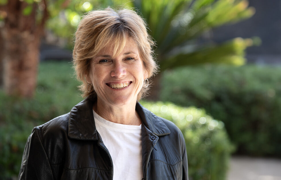 Portrait of Ellen Curtis in a black leather jacket and white shirt, smiling outdoors with blurred greenery behind