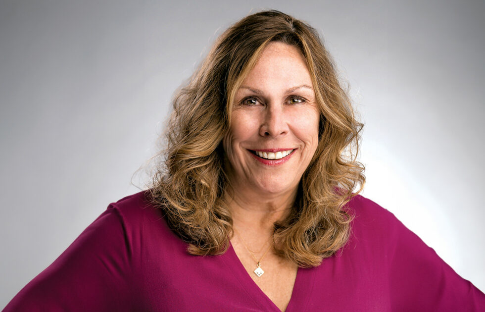 Geri LaChance in a magenta top smiling confidently with hands on hips against a gray background