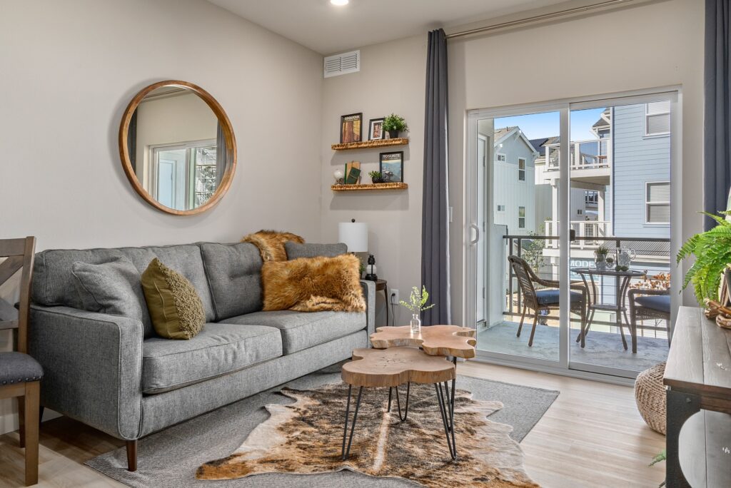 Modern apartment living room with gray couch, rustic decor, and sliding glass door opening to a balcony at Harvest Lofts.