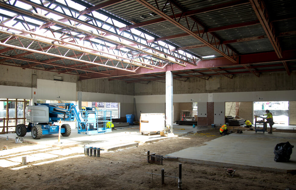 Interior view of an active construction site with workers and utility lifts under steel roof beams