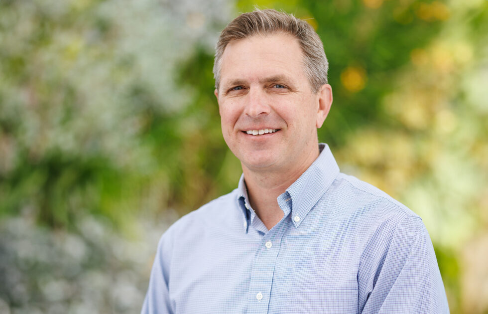 Headshot of Jim Dunning smiling in a light blue shirt with trees and greenery in the background