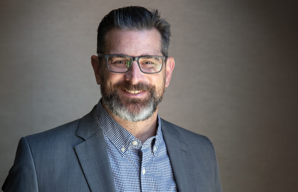 Joe Cleary smiling in a gray blazer and checkered shirt with glasses, standing in front of neutral wall