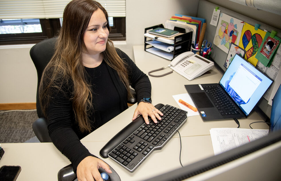 Maria Lopez-Pacheco working at her office desk, using a computer with colorful artwork on the wall