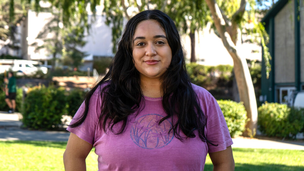 A confident student stands outdoors on a sunny day, smiling in front of a leafy campus backdrop wearing a purple t-shirt.