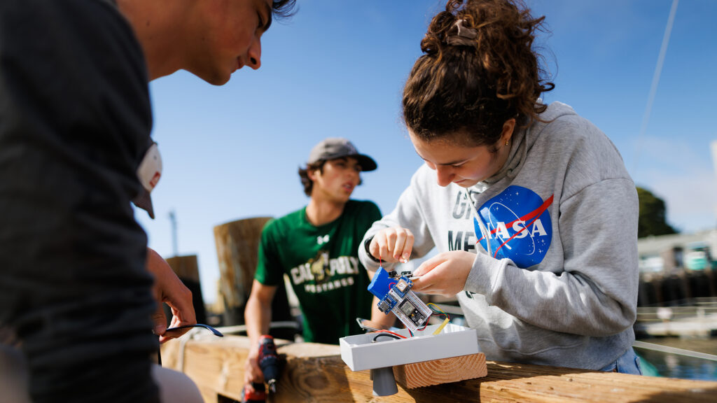 Students collaboratively work on assembling an electronic device outdoors, with one student in a NASA hoodie focused on connecting components while others observe and assist.