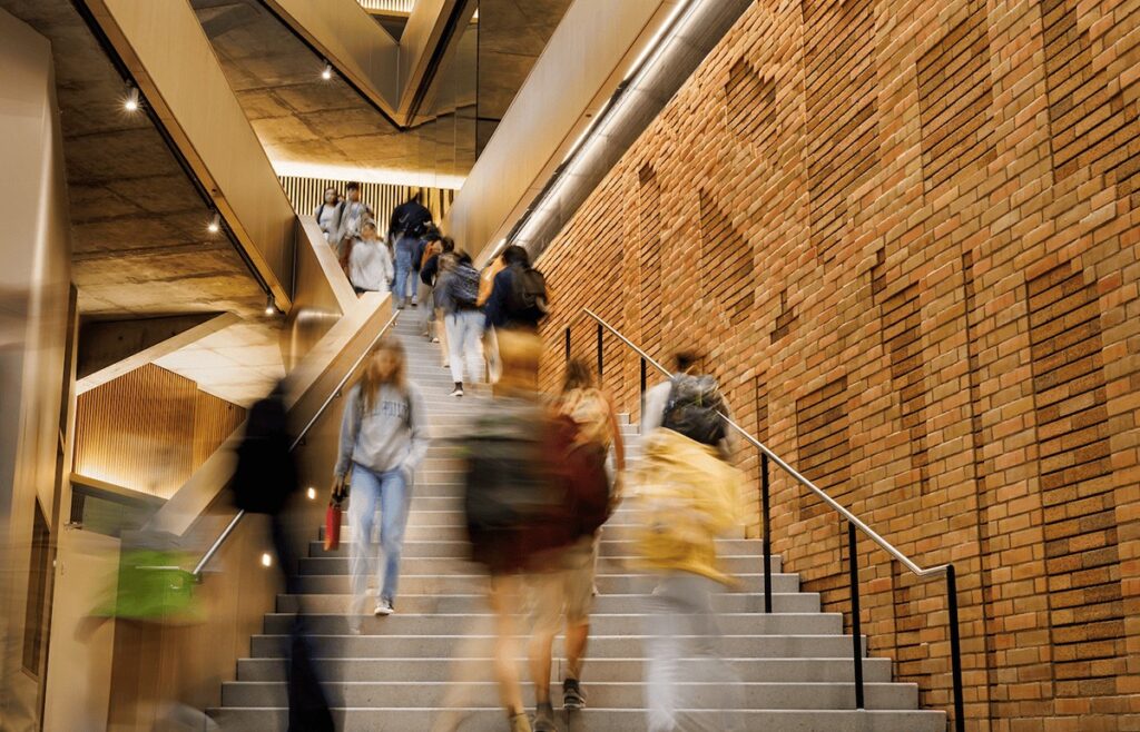 Students walk up and down a wide staircase inside a modern academic building with warm lighting and a geometric brick wall design, creating a dynamic, blurred motion effect.