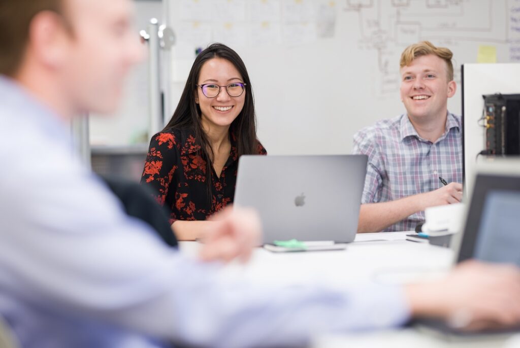 Three colleagues smiling and collaborating in a bright, modern office with laptops