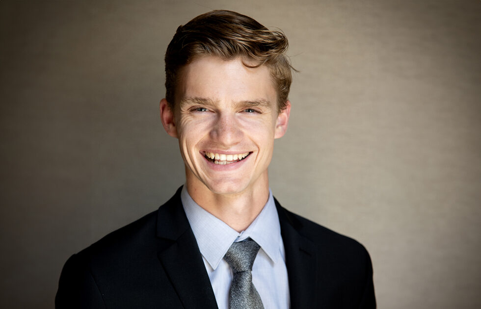 Toby Walpole smiling in a black suit and gray tie, standing in front of a neutral backdrop