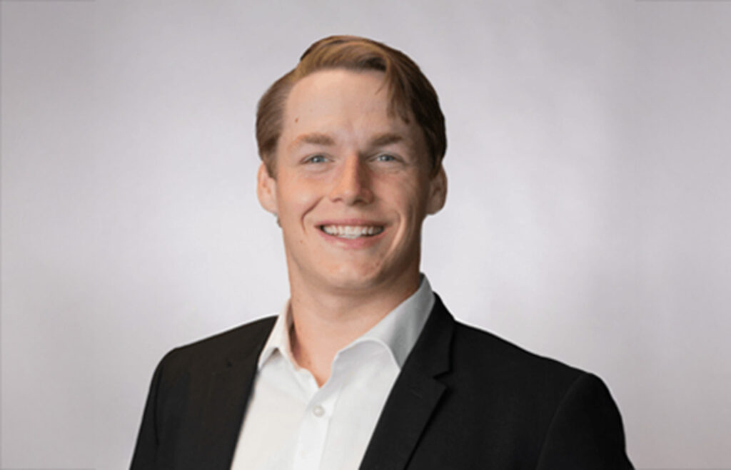 Professional headshot of a smiling person wearing a black suit jacket and white dress shirt against a light gray background.