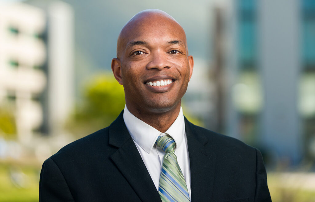 Professional headshot of a man in a black suit and striped green tie, smiling outdoors with blurred buildings in the background.