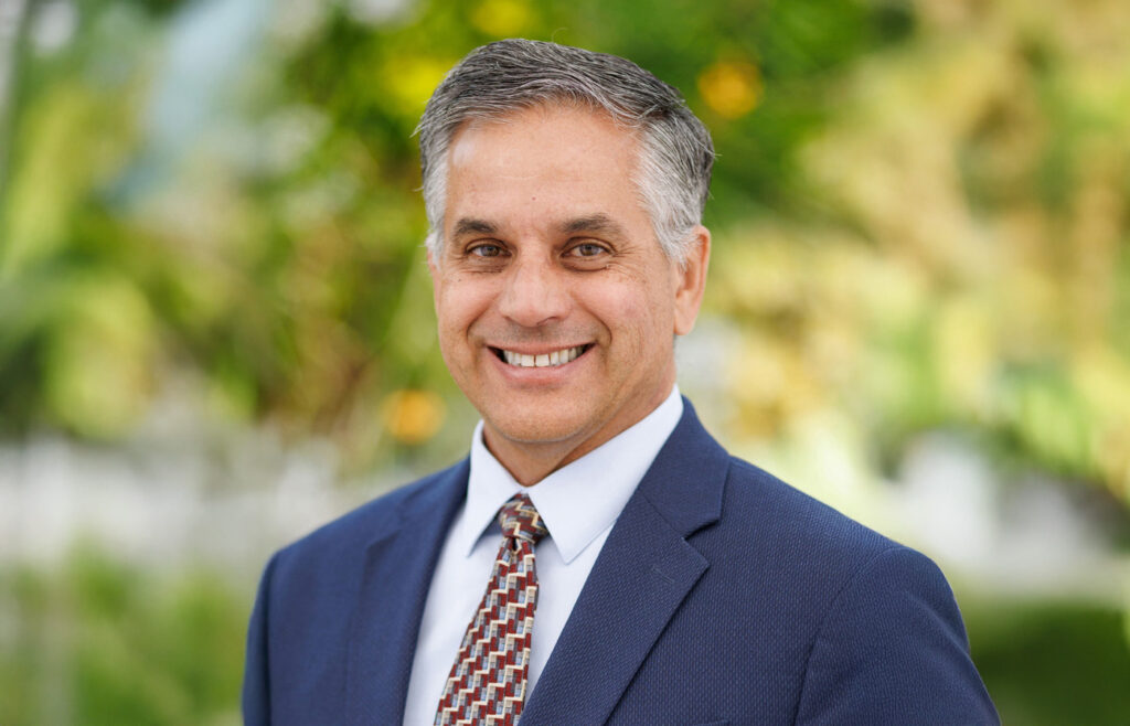 Professional headshot of a man in a navy suit and patterned tie, smiling outdoors with greenery in the background.
