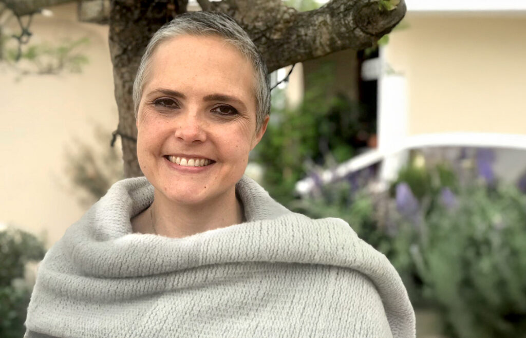 Smiling woman with short gray hair wearing a soft light gray knit shawl, standing outdoors near a tree with blurred greenery and a house in the background.