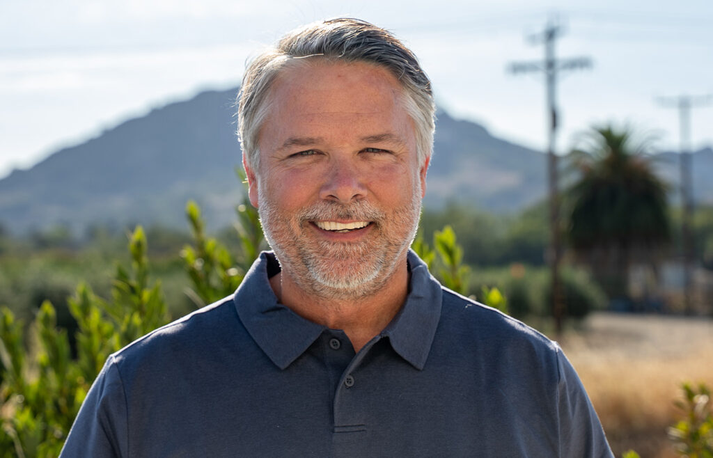 Portrait of a smiling middle-aged man with gray hair and beard standing outdoors in San Luis Obispo with a mountain in the background.