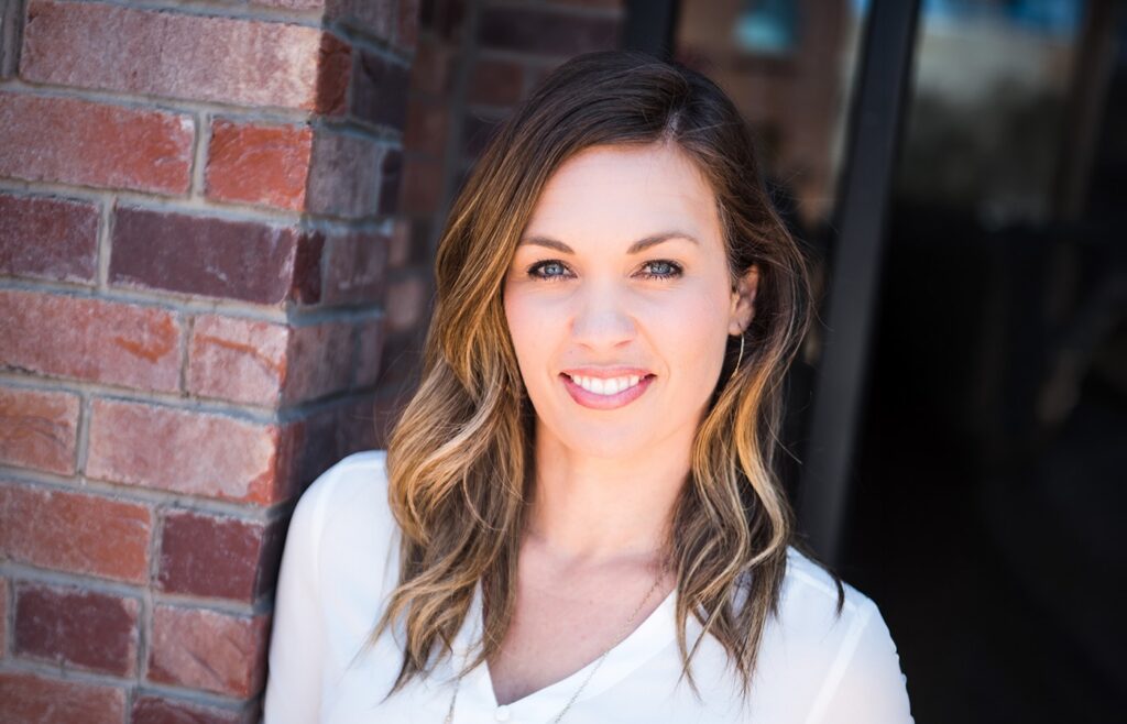 Professional headshot of a smiling woman with shoulder-length highlighted brown hair, standing against a red brick wall outdoors, wearing a white blouse and looking directly at the camera in natural light.