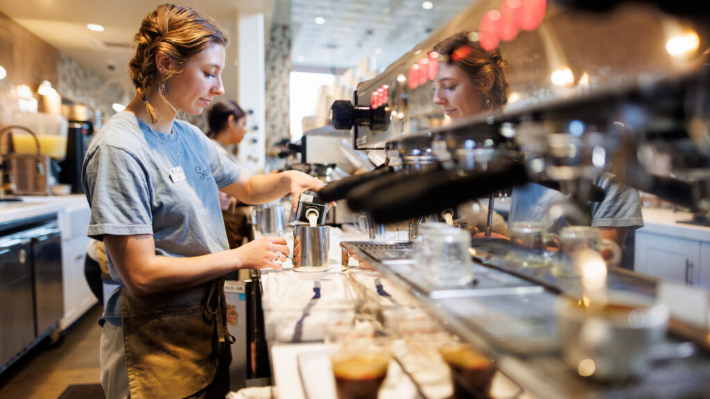 A barista pours milk into a metal pitcher beside an espresso machine.