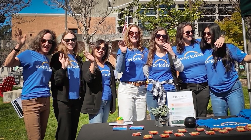 Seven Workday team members wave behind a Workday 101 information table at an outdoor campus event.