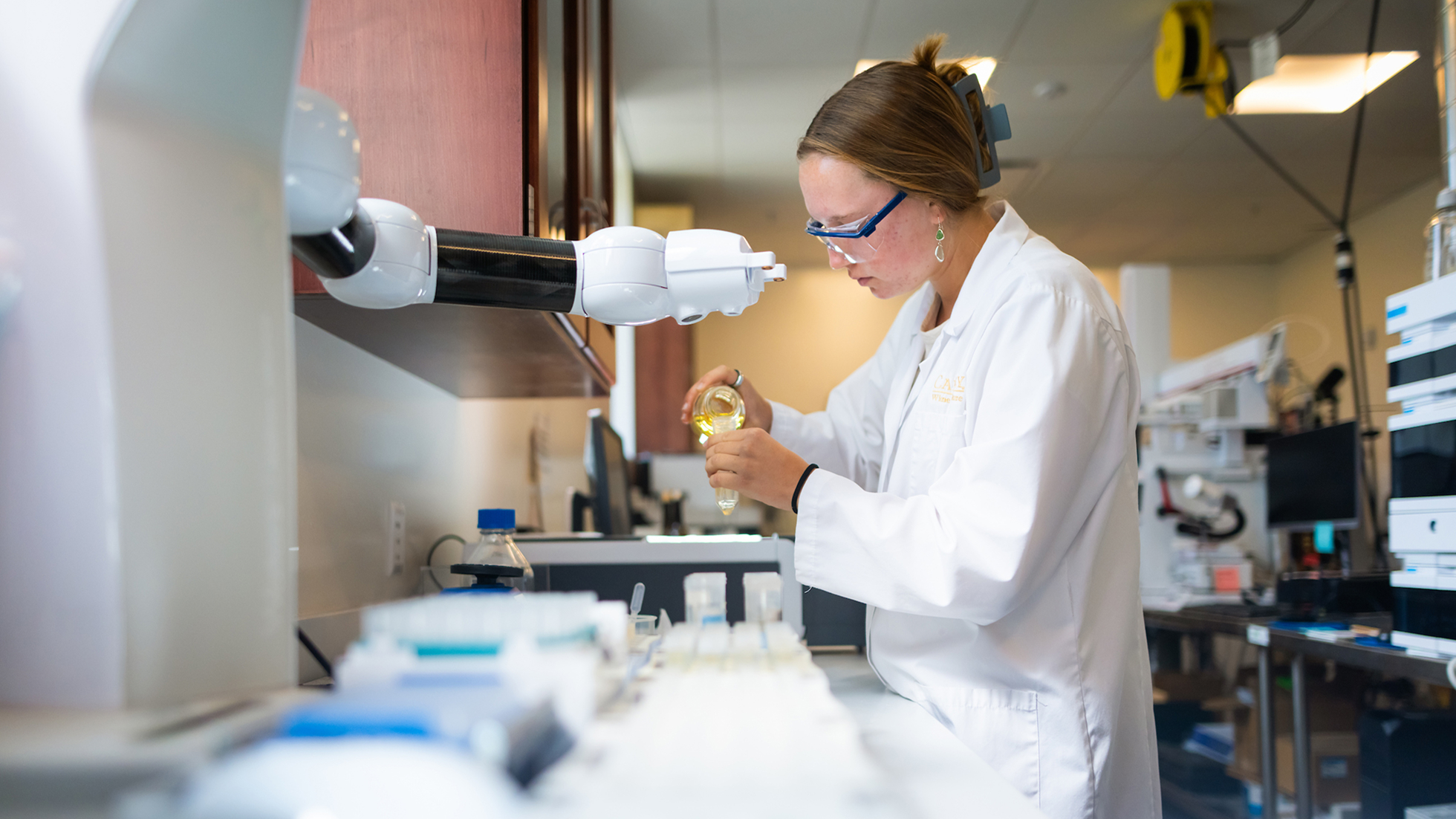 A researcher wearing safety glasses and a lab coat examines a yellow liquid sample in a laboratory.
