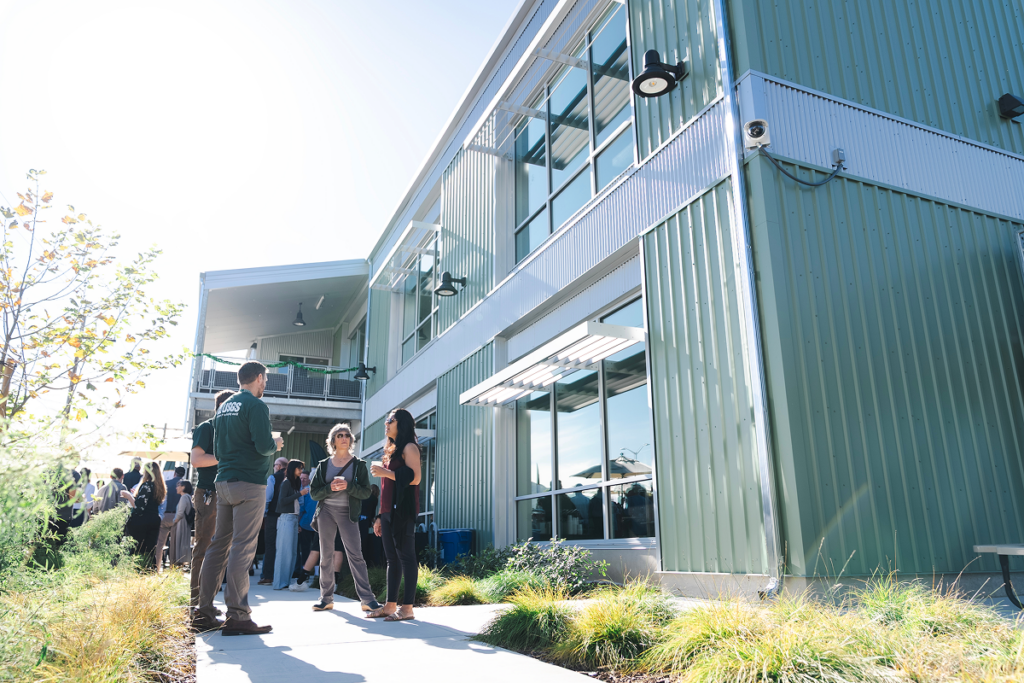 People converse outside a modern green metal building during a daytime gathering.