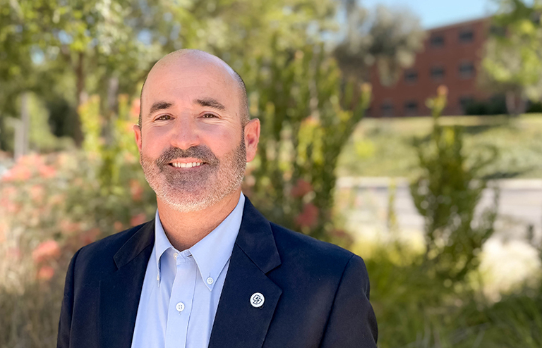 Professional headshot of Russell wearing a navy blazer and light blue shirt outdoors.