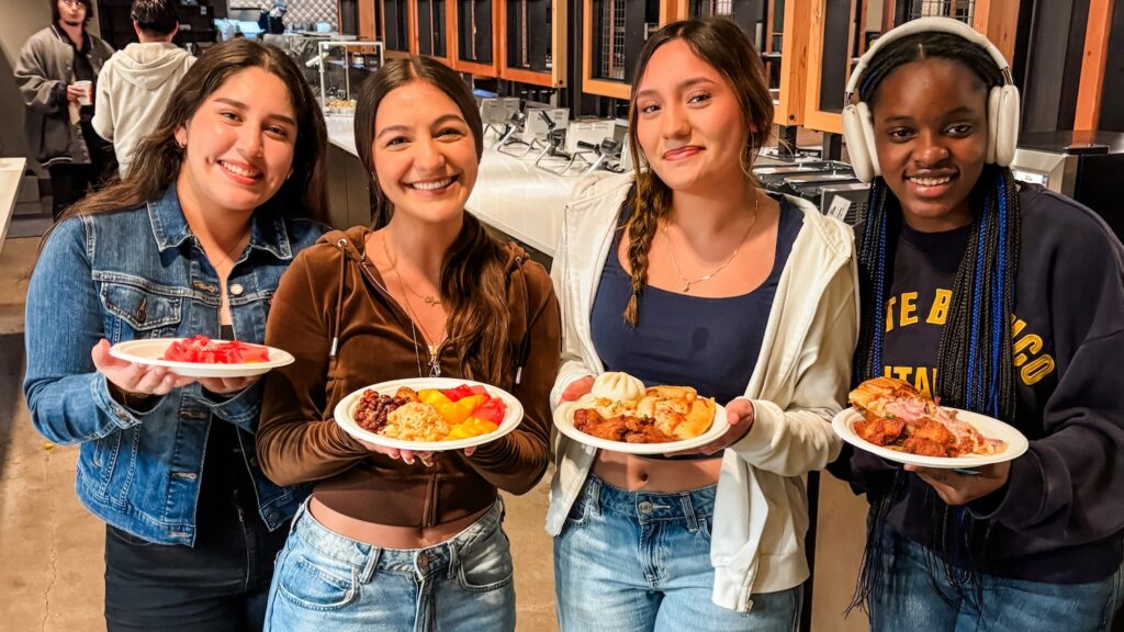 Four students smile while holding plates of food in a dining hall.